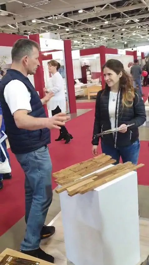 Exhibitor explaining interlocking teak wall panel samples on a white pedestal to a visitor holding a brochure, red carpet hall at HOMEDesign Budapest 2025