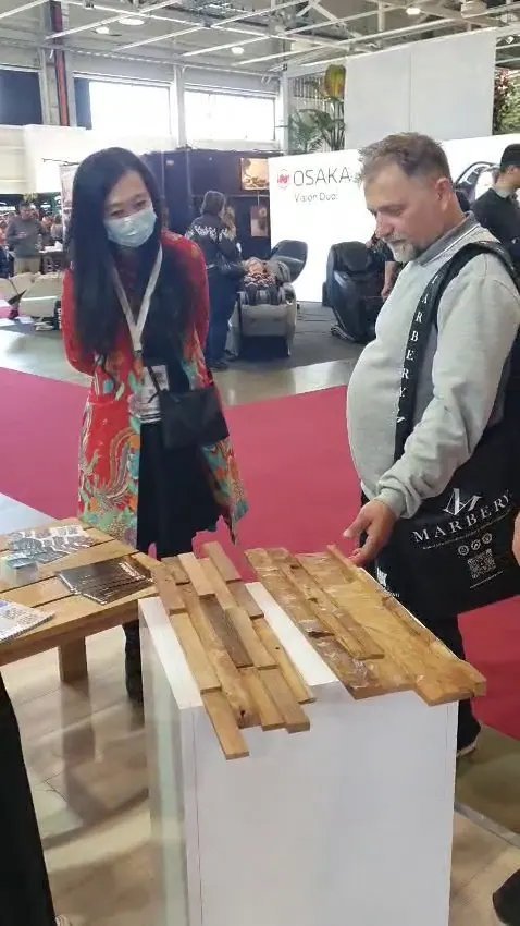 Buyer and exhibitor inspecting textured 3D teak wall cladding samples on a white pedestal at the Indonesia Pavilion, HOMEDesign Hungexpo 2025