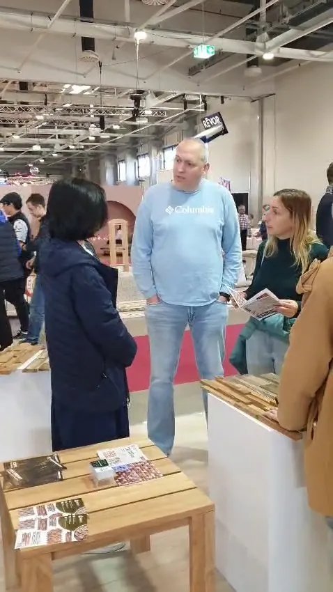 Trade visitors and staff gathered around teak wall panel samples and product brochures on a wooden table at HOMEDesign Budapest 2025