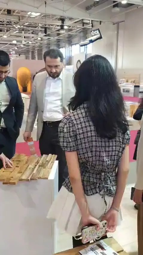 Visitors examining light brown teak wall cladding samples on a white display pedestal at HOMEDesign Budapest 2025