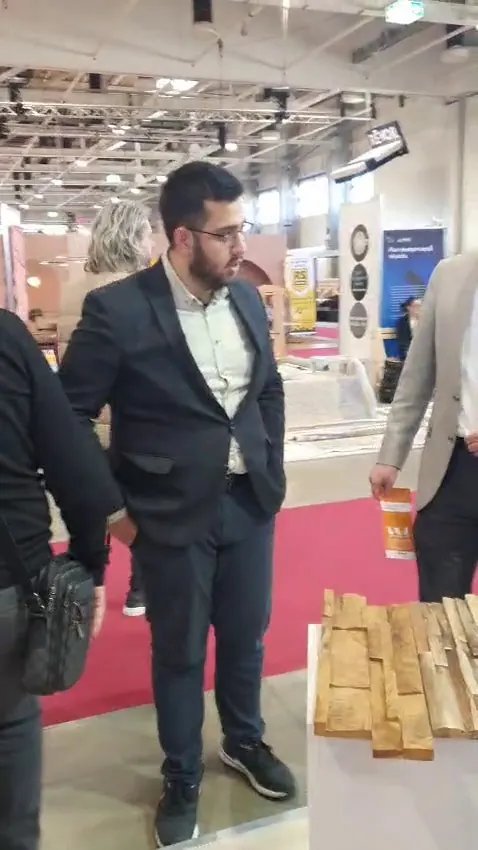 Trade visitor in a dark suit examining a textured teak wall panel sample on a white pedestal, red carpet aisle at Hungexpo Budapest