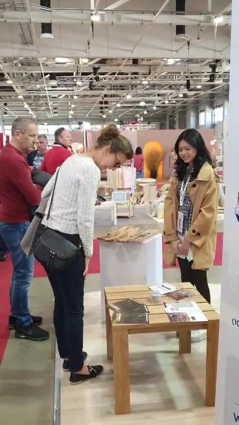 Visitor inspecting teak wall cladding samples on a white pedestal while booth staff smiles, brochures on a wooden table, HOMEDesign Budapest 2025