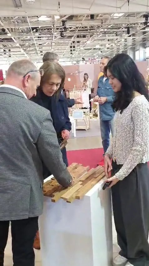 Prospective buyers examining teak wood wall cladding samples on a white pedestal with booth staff at HOMEDesign Budapest 2025, Hungexpo