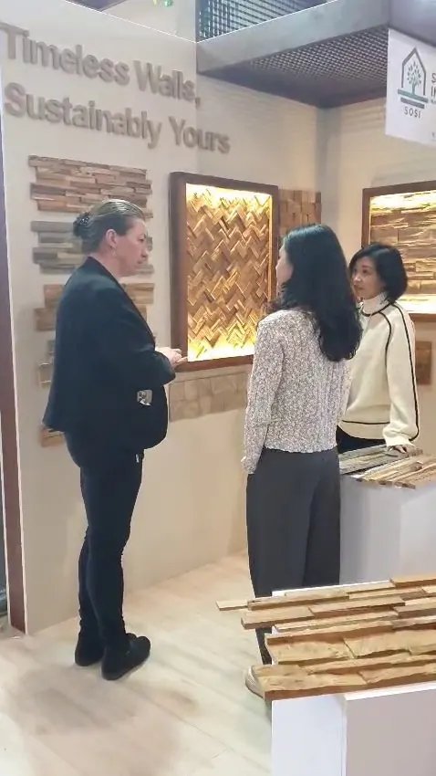 Exhibitor talking with visitors beside wall-mounted teak and bamboo panel samples and a backlit herringbone wood display, HOMEDesign Budapest 2025