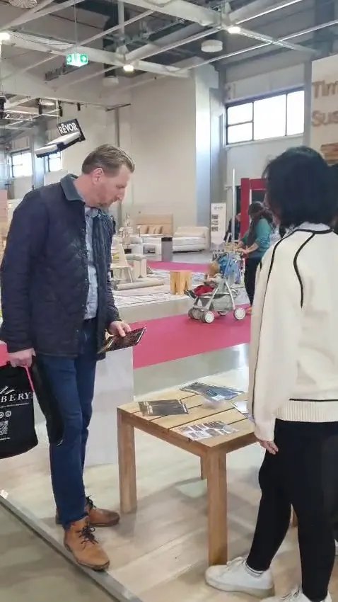 Visitor and exhibitor reviewing product brochures on a wooden sample table at the SOSI booth, HOMEDesign exhibition hall with red carpet, Hungexpo Budapest
