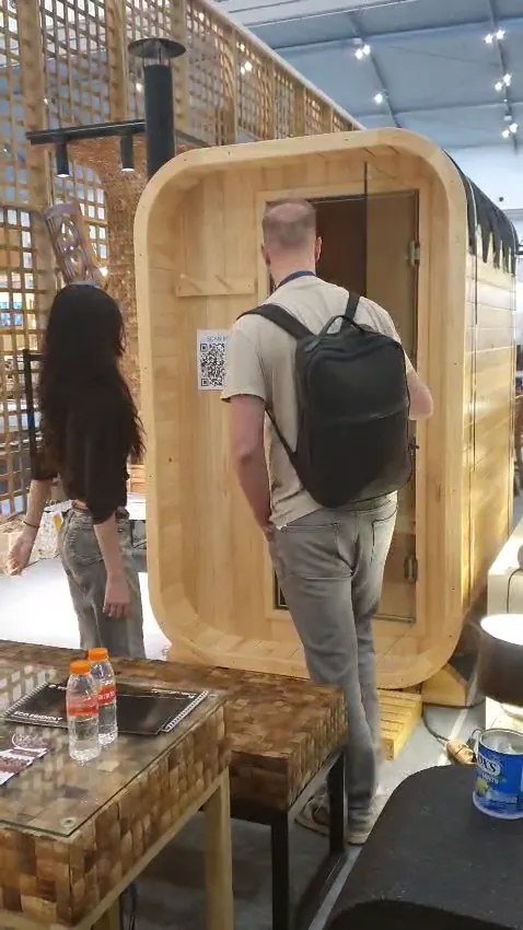 Visitor inspecting a vertical barrel pinewood sauna with QR sticker at the SOSI IFEX booth, teak mosaic table in the foreground