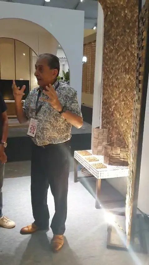 Exhibitor gesturing beside illuminated teak mosaic wall panels and white mosaic sample benches at the IFEX booth