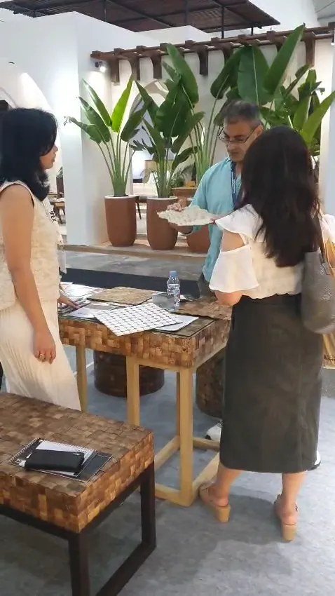 SOSI staff presenting a white mosaic wall panel sample to a visitor at a teak mosaic display table, IFEX booth with tropical planters