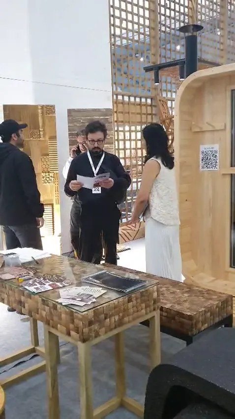 Buyers and staff around teak mosaic furniture with brochures, barrel pinewood sauna and SCAN ME QR sticker visible at the SOSI IFEX booth