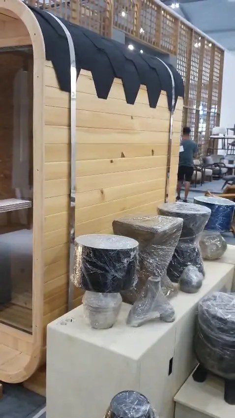 Vertical barrel pinewood sauna with black shingle roof and chrome bands beside mosaic furniture samples on a white counter at IFEX